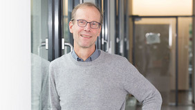 A man with short light brown hair and glasses leans smiling against a glass wall in a modern hallway. He is wearing a gray sweater over a shirt and a wristwatch. Glass doors and a bright interior can be seen in the background.