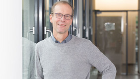 Ein Mann mit kurzen hellbraunen Haaren und Brille lehnt lächelnd an einer Glaswand in einem modernen Flur. Er trägt einen grauen Pullover über einem Hemd und eine Armbanduhr. Im Hintergrund sind Glastüren und ein heller Innenraum zu sehen.