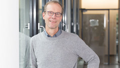 Ein Mann mit kurzen hellbraunen Haaren und Brille lehnt lächelnd an einer Glaswand in einem modernen Flur. Er trägt einen grauen Pullover über einem Hemd und eine Armbanduhr. Im Hintergrund sind Glastüren und ein heller Innenraum zu sehen.