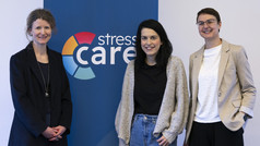 Drei Frauen stehen lächelnd vor einem Roll-up-Banner mit der Aufschrift „stress care“ und der Website www.stress-care.de . In der Mitte ist das farbige Logo von Stress Care zu sehen. Die Personen sind in Business- und Freizeitkleidung gekleidet und stehen nebeneinander in einem hellen Innenraum.