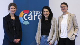 Drei Frauen stehen lächelnd vor einem Roll-up-Banner mit der Aufschrift „stress care“ und der Website www.stress-care.de . In der Mitte ist das farbige Logo von Stress Care zu sehen. Die Personen sind in Business- und Freizeitkleidung gekleidet und stehen nebeneinander in einem hellen Innenraum.
