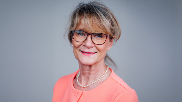 Mittelalte blode Frau in lachsfarbener Bluse vor einem grauen Hintergrund.