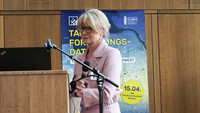 A middle-aged blonde woman stands at a lectern with a microphone.