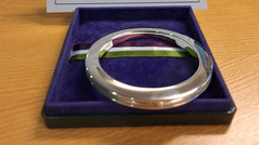 A polished silver bangle bracelet displayed in a purple velvet-lined box on a wooden table, with a multicolored ribbon beneath it.