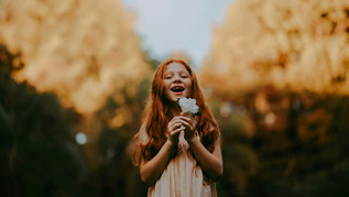 A cheerful girl with long red hair stands outdoors, holding a white flower in her hands and laughing. In the background, autumnal golden trees and a blue sky can be seen.