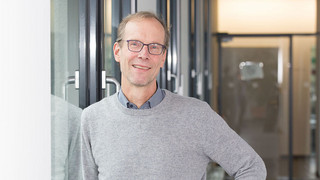 Ein mittelalter Mann steht an eine Wandgelehnt vor einem Fenster. 