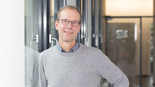 Ein mittelalter Mann steht an eine Wandgelehnt vor einem Fenster. 